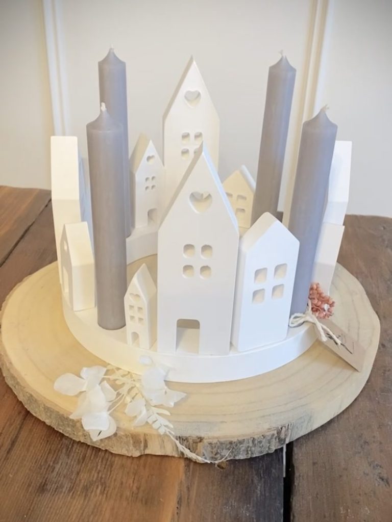 Dekorativer Adventskranz mit weißen Gebäuden und grauen Kerzen auf einer Holzplatte.