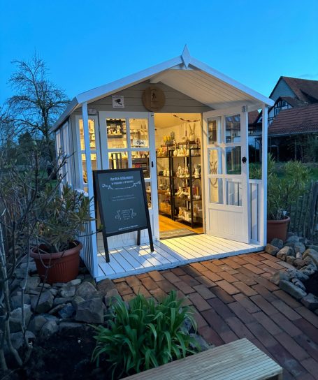 Ein beleuchtetes Gartenhaus mit Glasfenstern und einem schwarzen Schild an der Vorderseite.