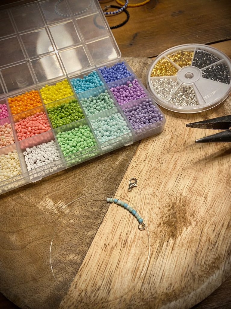 Bunte Perlen in einer Box, Werkzeuge und ein Holzbrett im Hintergrund.