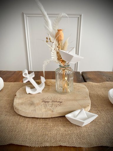 Holzbrett mit weißen Papierschiffchen, Ankern und Glasvase mit Trockenblumen